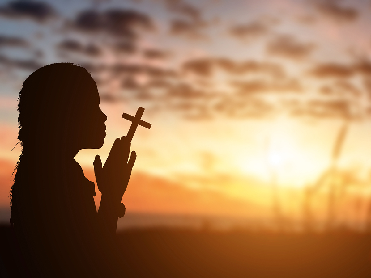 Silhouette of little girl holding christian cross in hands to praying for blessing from god at sunset background.