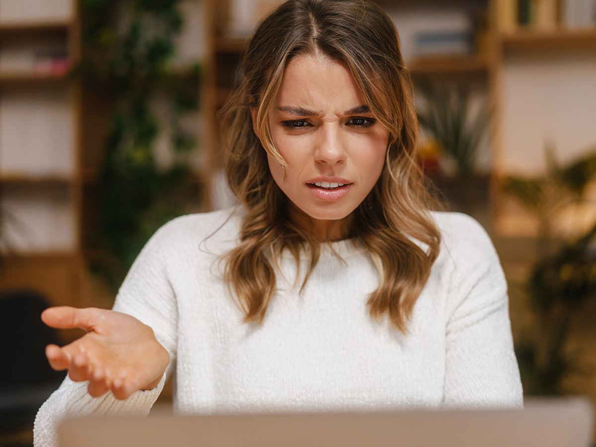 Frustrated confused young woman manager looking at laptop computer while sitting at the office desk, gesturing