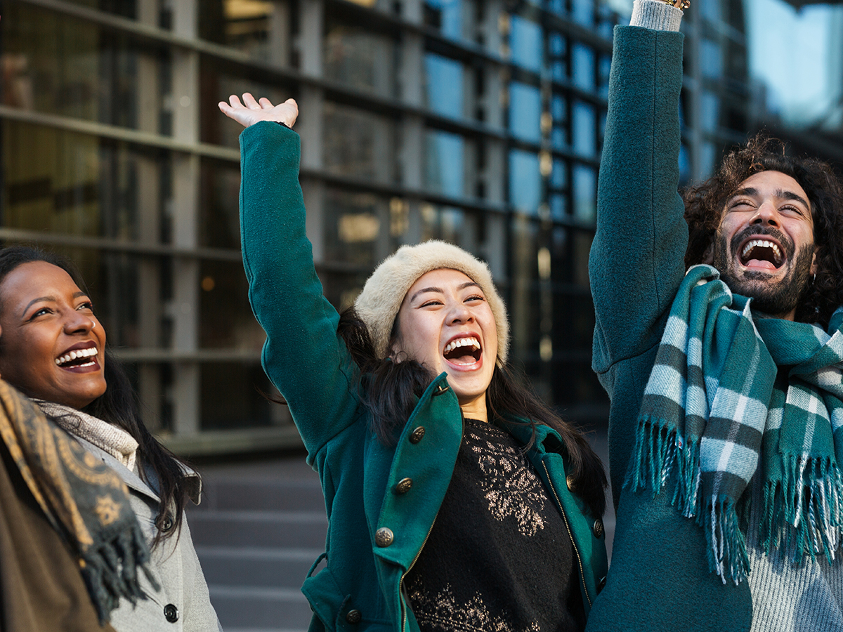 Cheerful diverse man and women in outerwear raising arms and laughing after stacking hands while celebrating success on street together