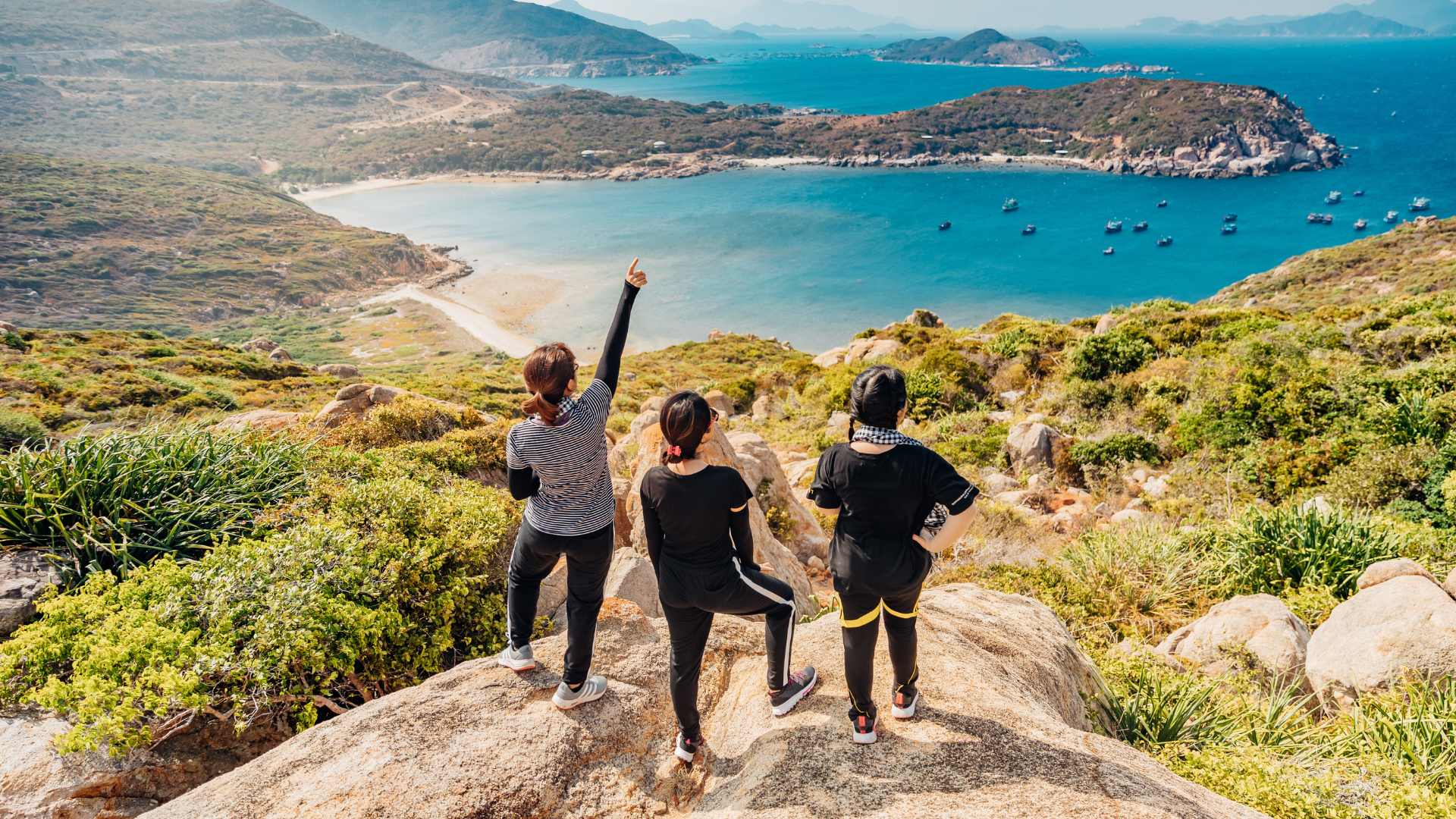 three young women celebrate reaching the top of a mountain, overlooking a beautiful coastline on a sunny day